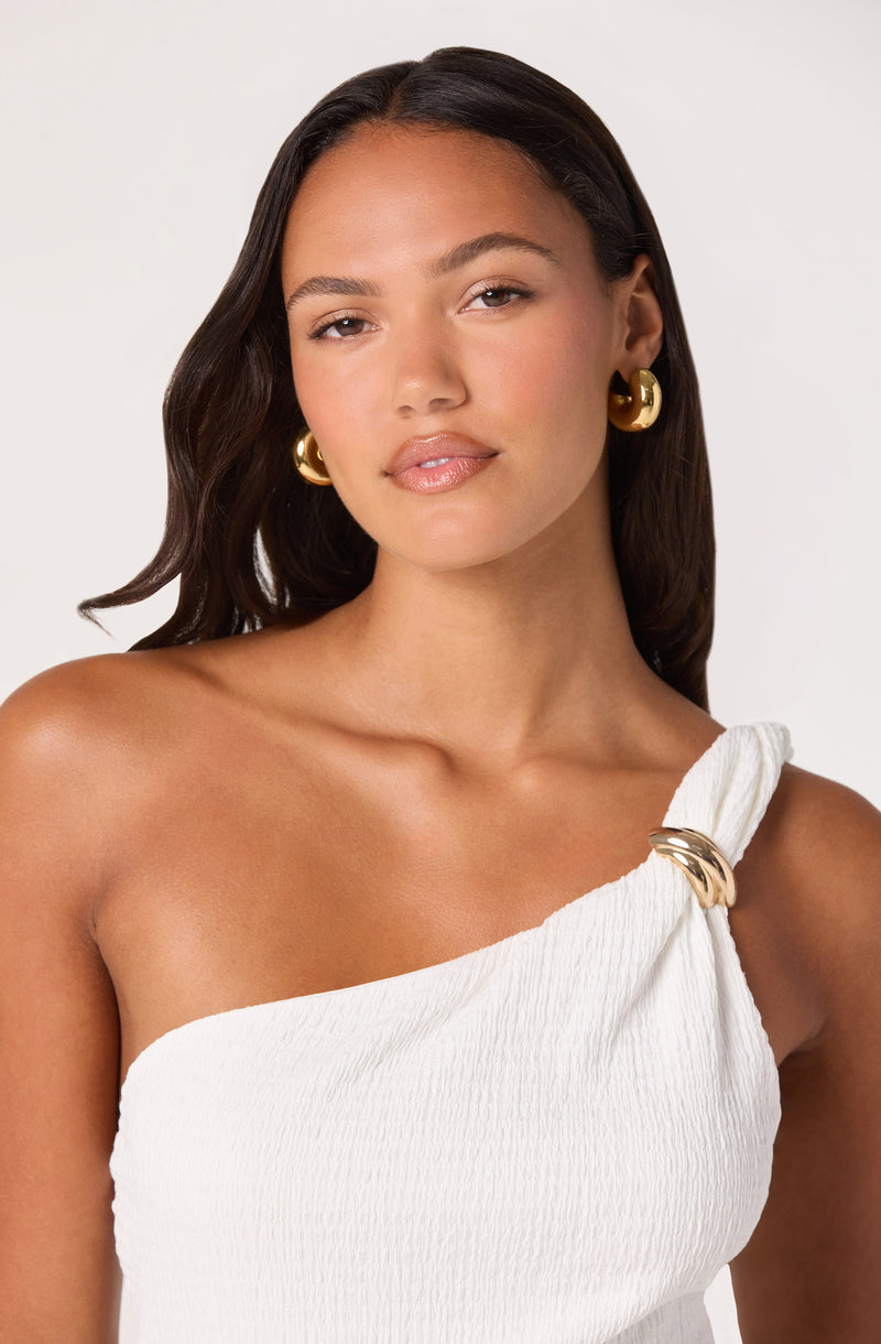 Woman wearing a white, textured, one-shoulder top with a gold embellishment at the shoulder.