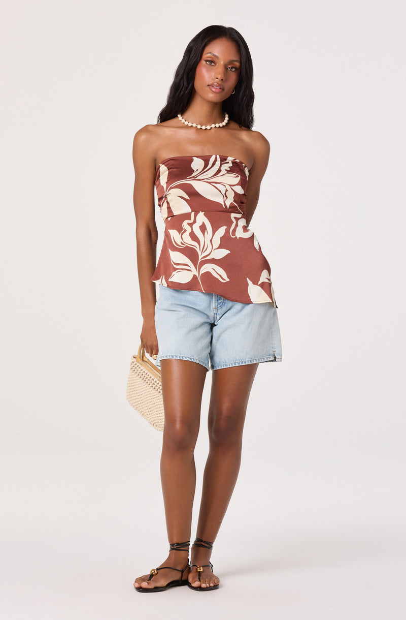 Woman in a strapless, brown and cream floral top with light blue denim shorts and black sandals.