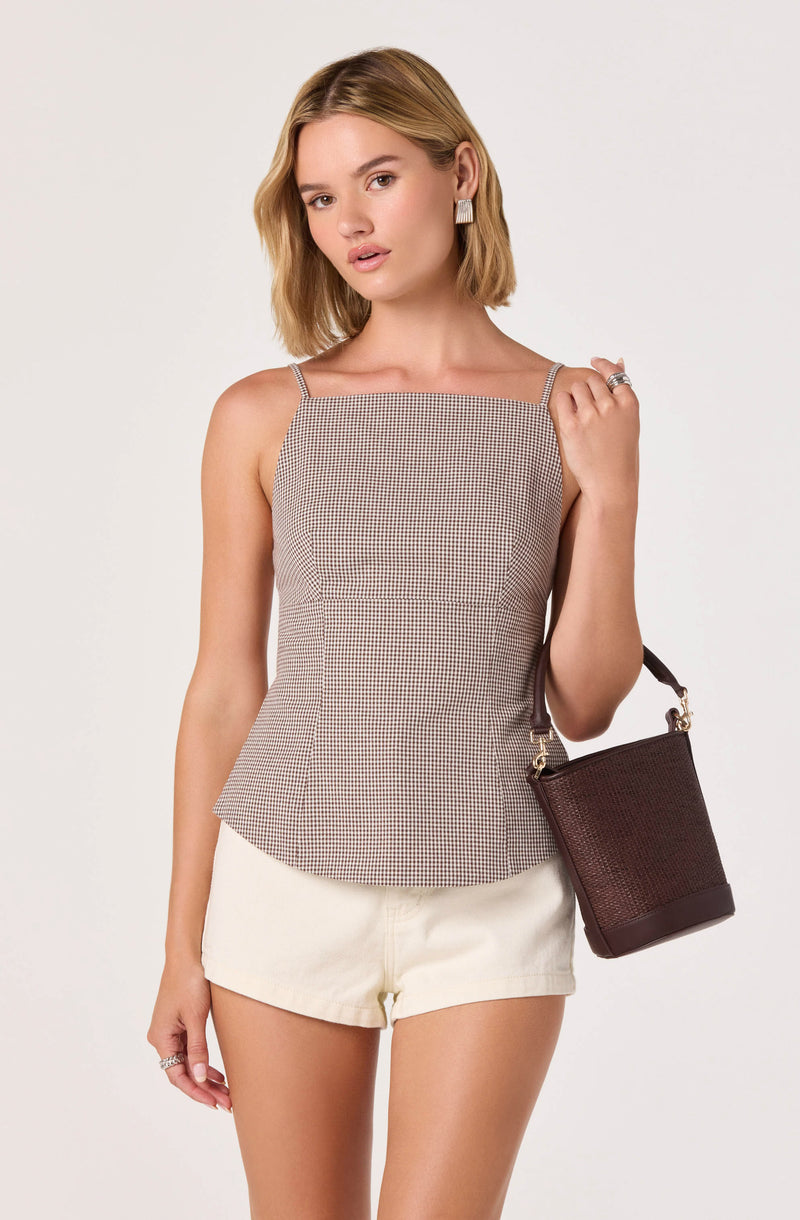 Woman wearing a sleeveless checkered top with thin straps and cream shorts, holding a dark brown handbag.