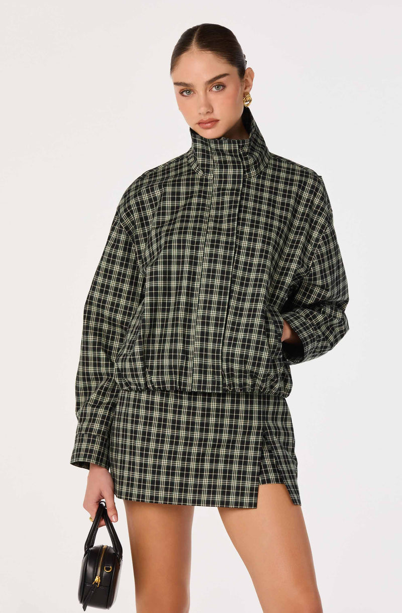 Woman wearing a black and green plaid jacket with a matching skirt and gold earrings, holding a black handbag.