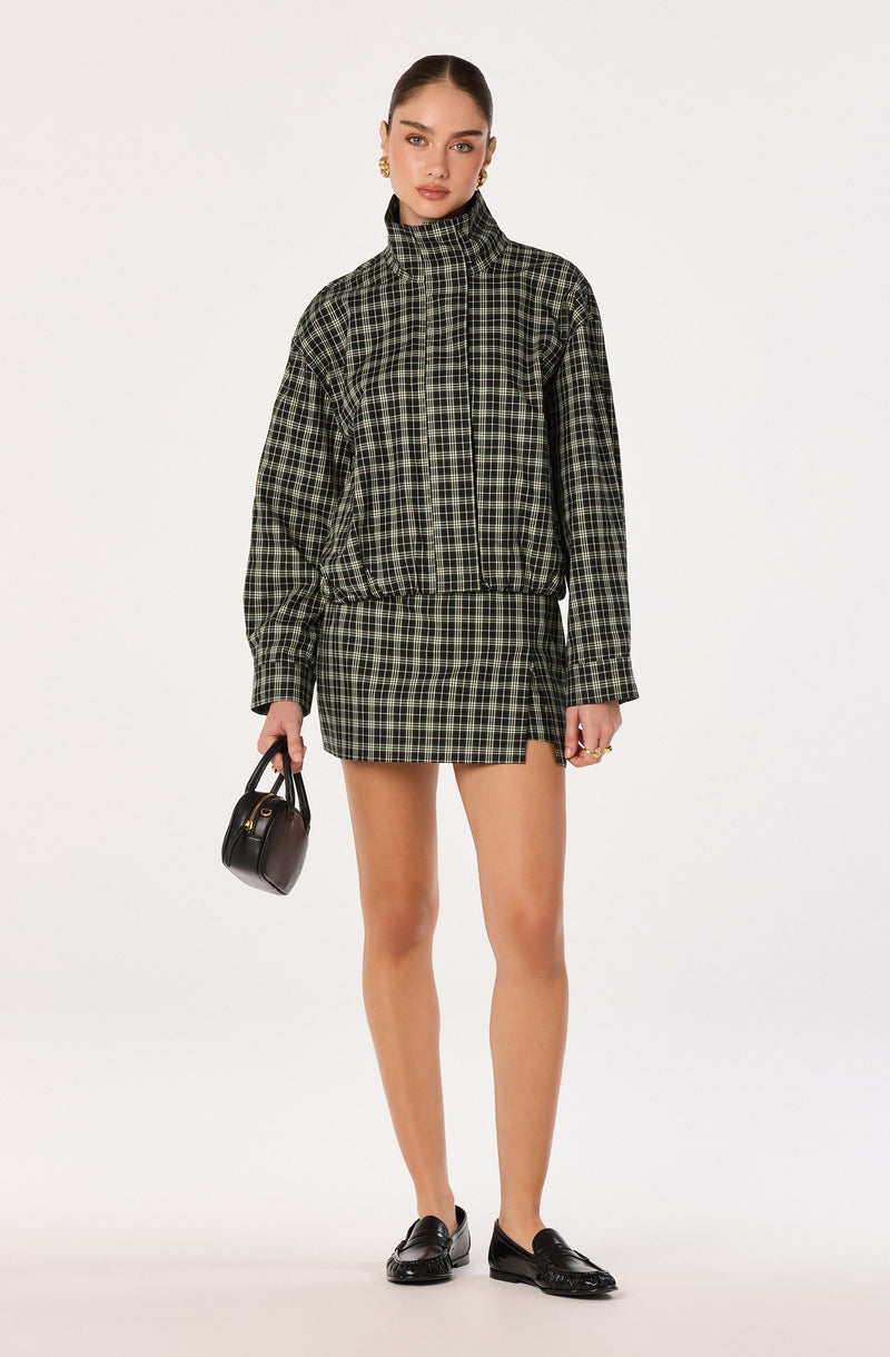 Model wears a black and green plaid jacket and matching skirt, with black shoes and handbag.