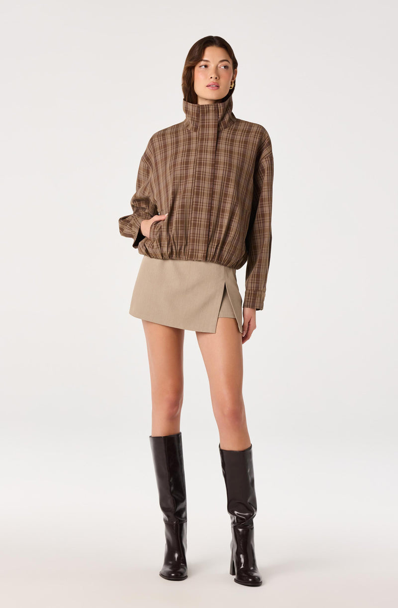 Woman wearing a brown plaid high-neck jacket, beige mini skirt with a slit, and black knee-high boots.