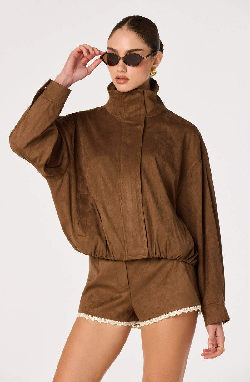 Woman in brown suede jacket and shorts with lace trim, wearing sunglasses.