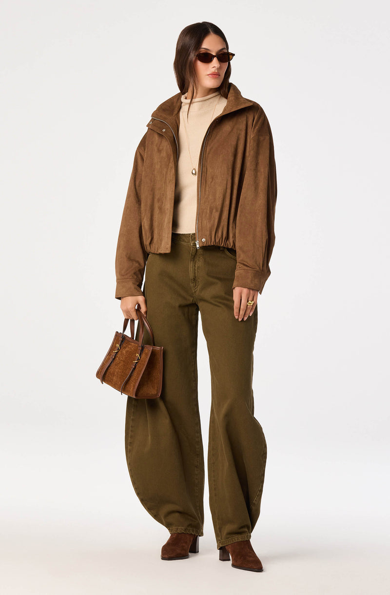 Woman wearing a brown suede jacket, beige top, olive green wide-leg pants, brown boots, and dark sunglasses, holding a brown handbag.