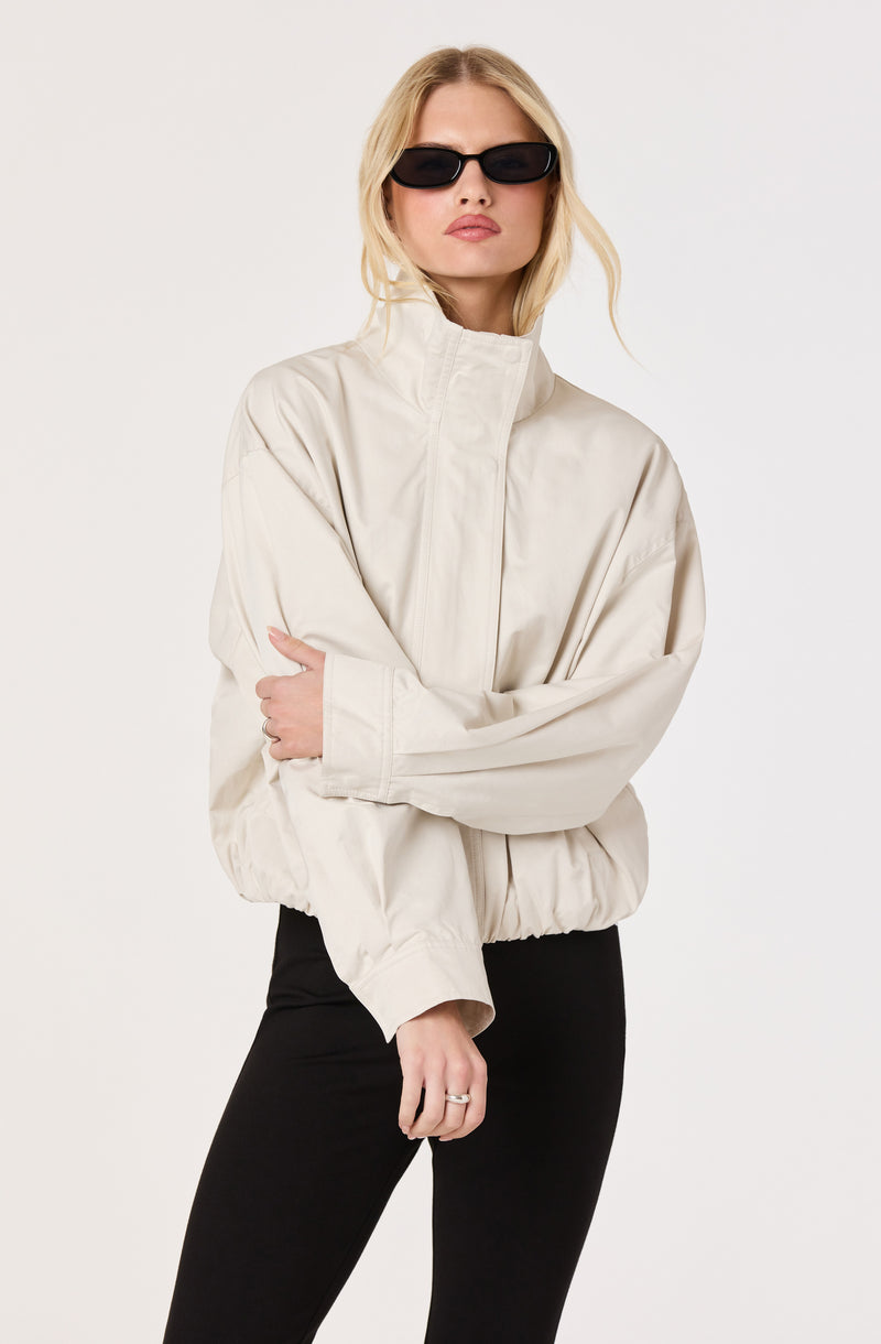 Woman in a beige oversized jacket and black pants, wearing black sunglasses.