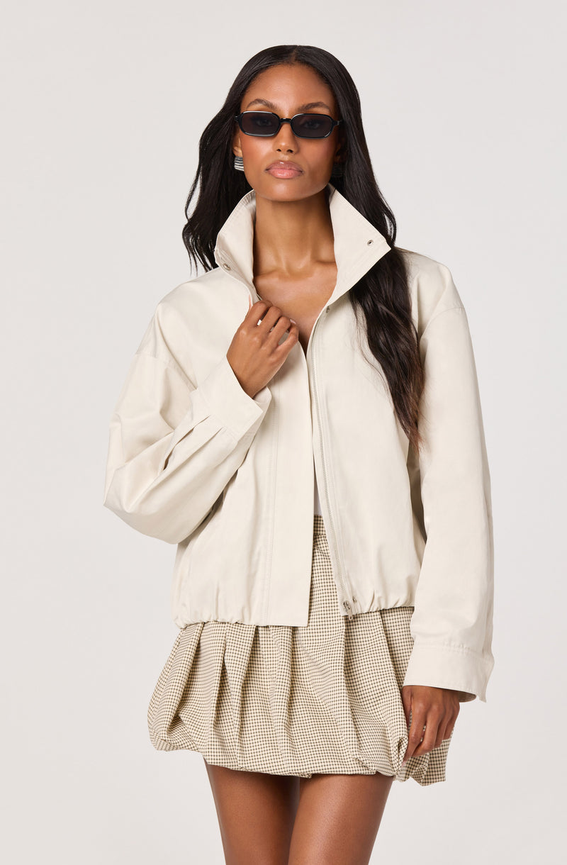 Woman in cream jacket with high collar, front zipper, and elastic hem, paired with a beige checkered skirt.
