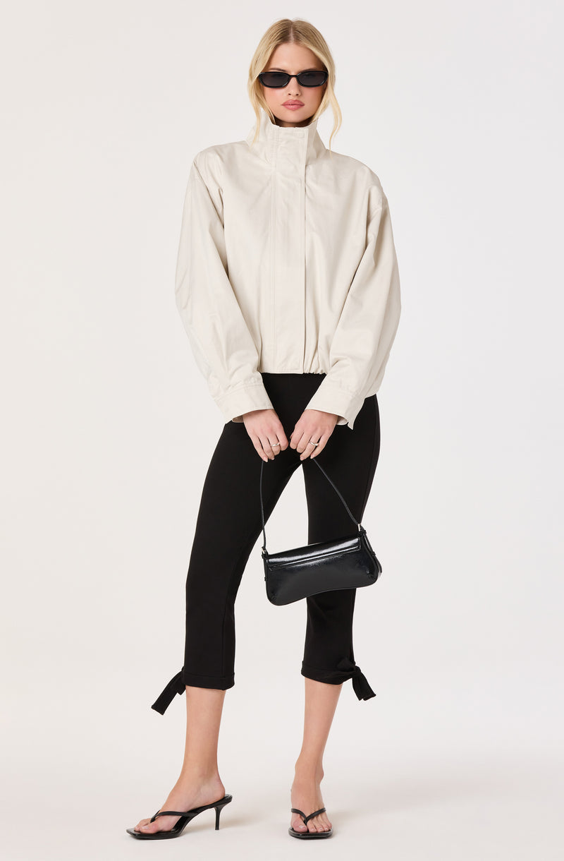 Woman wearing a beige high-neck jacket, black cropped pants with tied hems, black heels, and sunglasses, holding a black handbag.