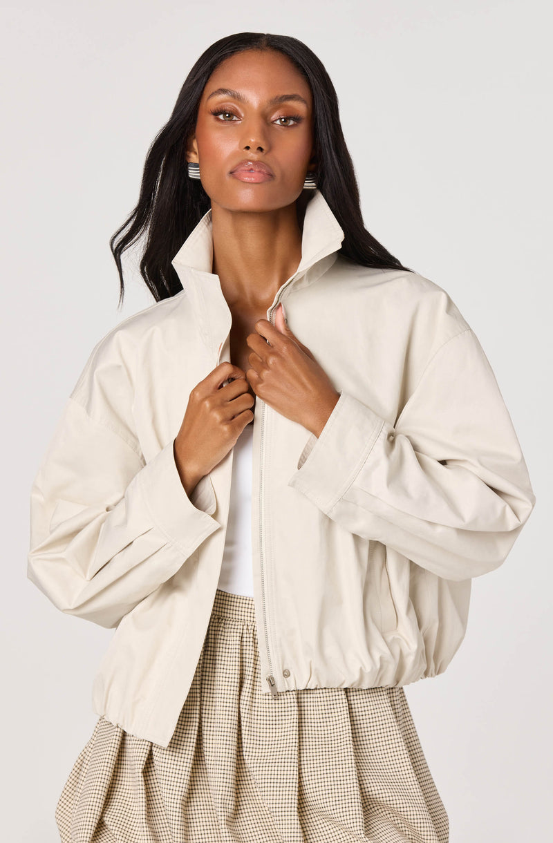 Woman in a cream, high-collared zip-up jacket and beige houndstooth skirt.