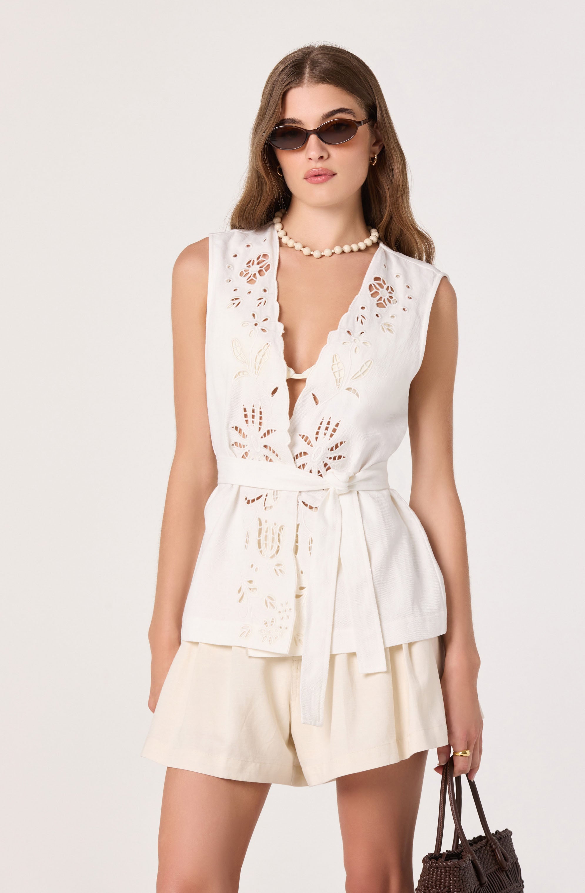 Woman in white sleeveless top with laser-cut floral details, beige shorts, and sunglasses.