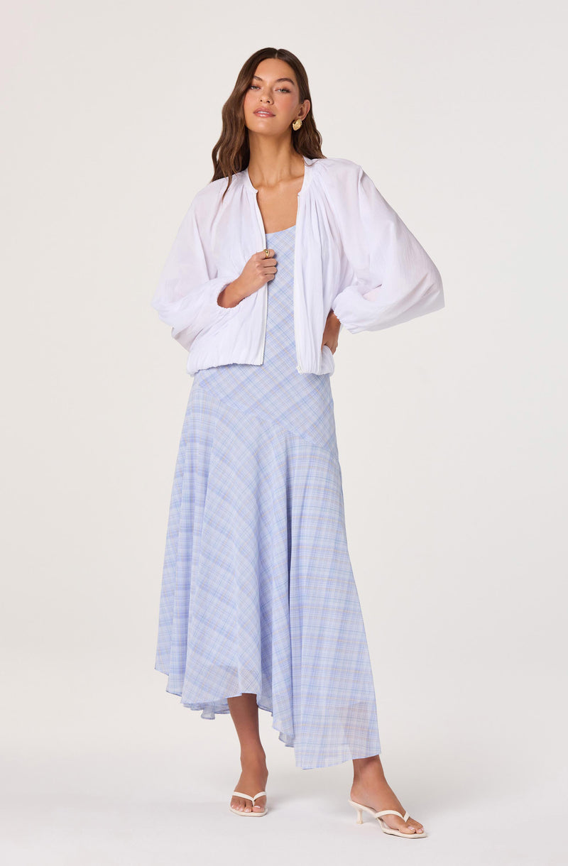 Woman wearing a white, long-sleeved, lightweight jacket over a light blue plaid dress, paired with white heels.