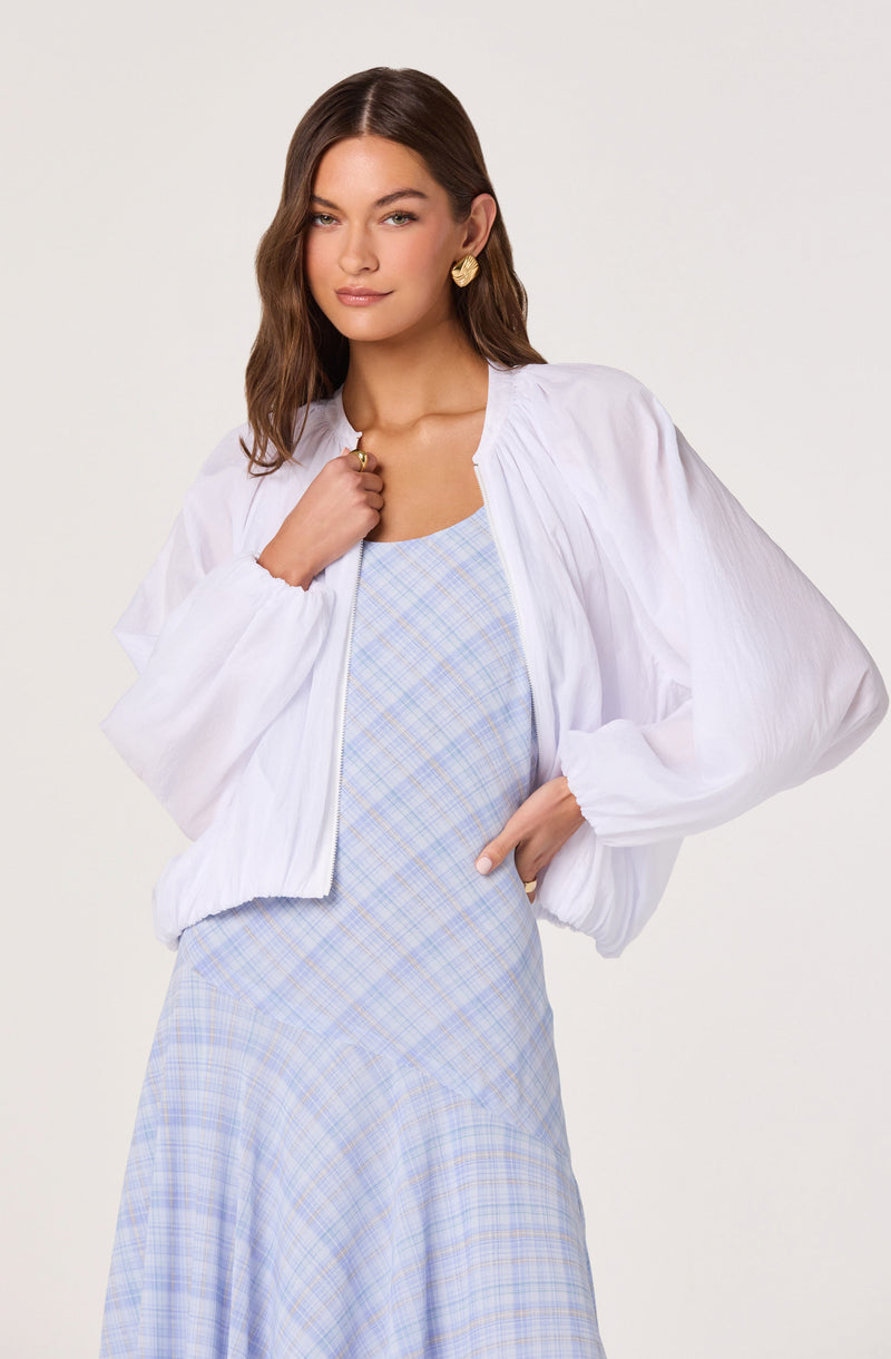 Woman wearing a white, long-sleeved, zip-up jacket over a light blue plaid dress.
