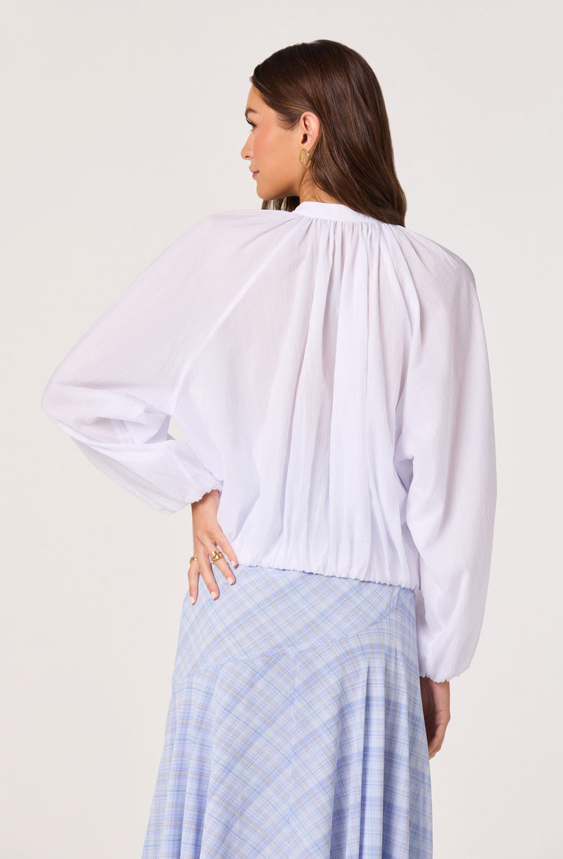 Woman wearing a white, long-sleeve, gathered blouse with an elastic hem and puffed sleeves, paired with a light blue plaid skirt.