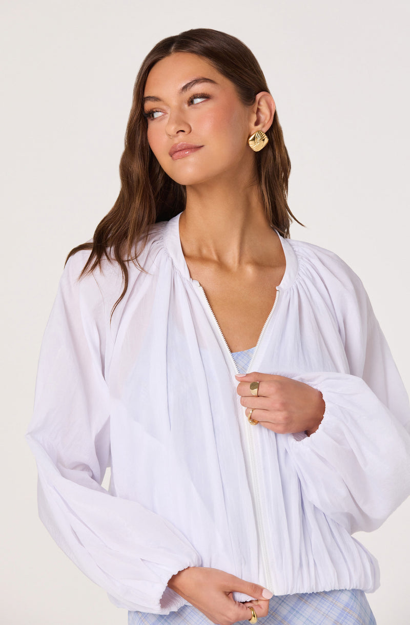 Woman wearing a white, zip-up, puff-sleeve jacket with gold earrings and rings.