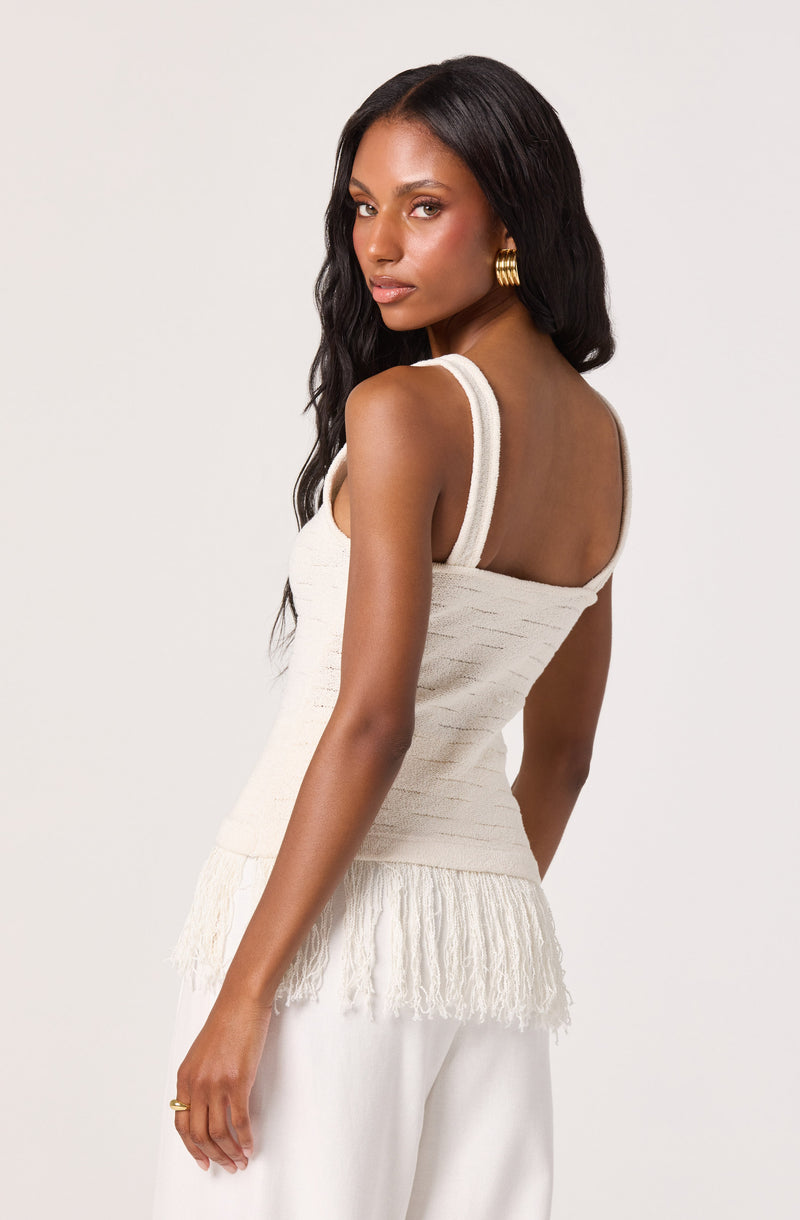 Woman wearing a sleeveless cream knit top with fringe at the hem and white loose pants, gold hoop earrings.