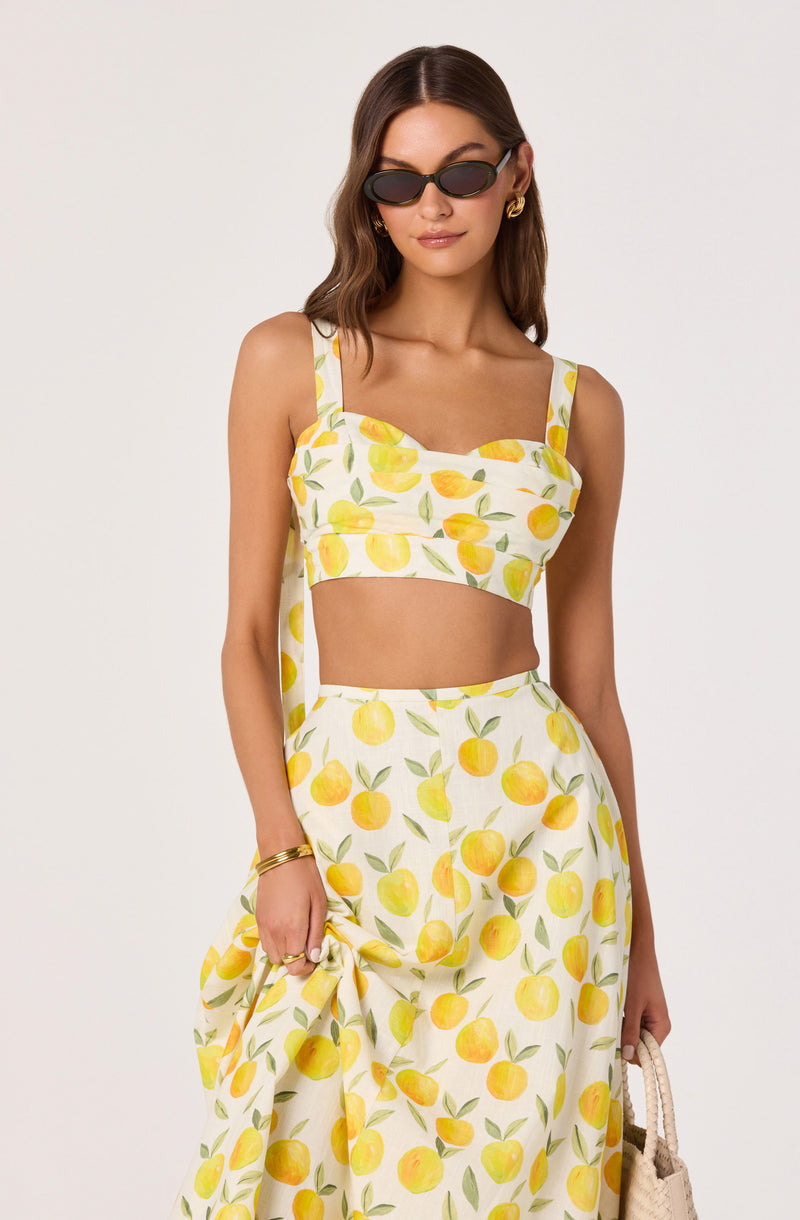 Woman wearing a lemon print crop top and matching flowy skirt, with dark sunglasses and gold accessories.