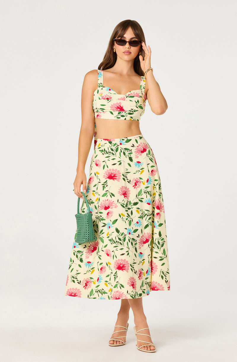 Woman wearing a floral crop top and matching long skirt with pink and green flowers, holding a green handbag, and wearing sunglasses.