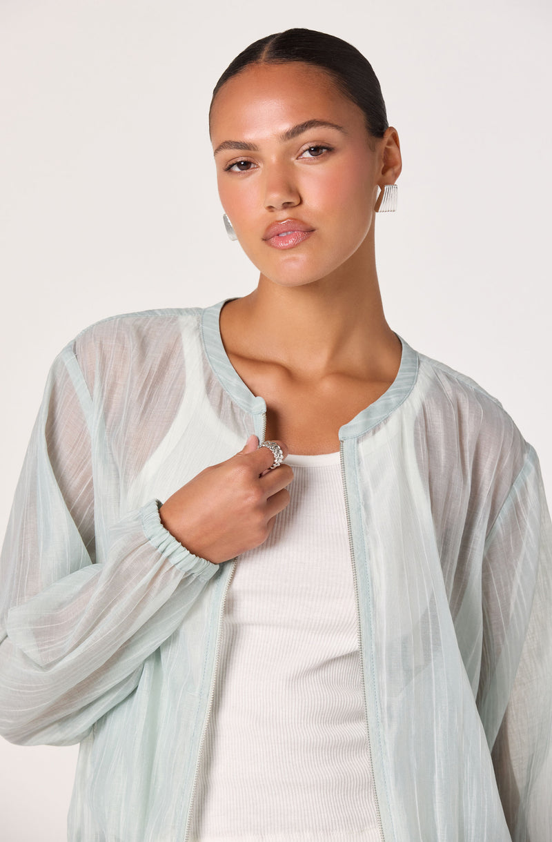 Woman wearing a sheer, light-colored jacket over a white top, with silver earrings and rings.