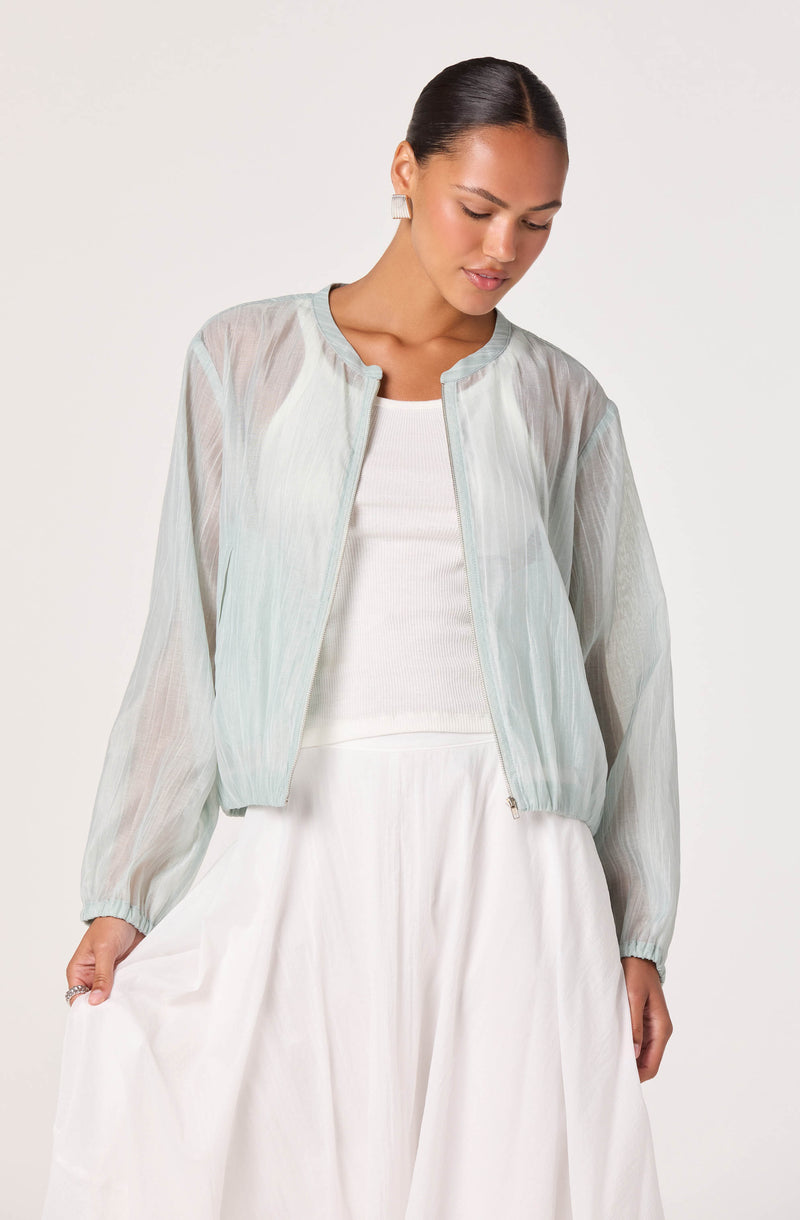 Woman wearing a light, semi-transparent, pastel-colored jacket over a white top and white skirt.