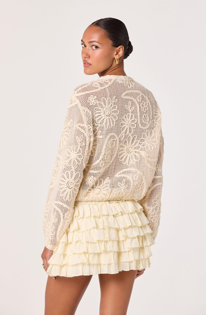 Woman wearing a cream crochet sweater with floral patterns and a ruffled cream skirt.
