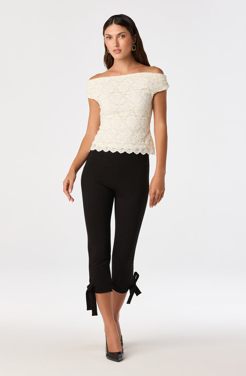Woman wearing an off-the-shoulder cream lace top and black cropped pants with tied bows at the hem.