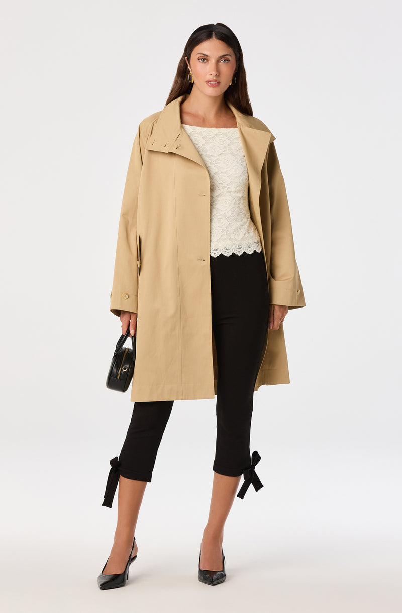 Woman wearing a beige trench coat, white lace top, black ankle-length pants with bows, and black high heels.