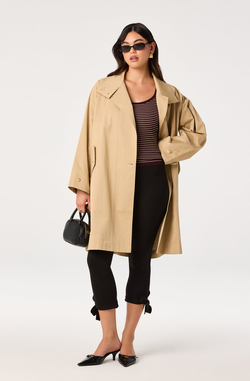 Woman in beige trench coat, black striped top, black cropped pants with bows, black heels, black purse, and sunglasses.
