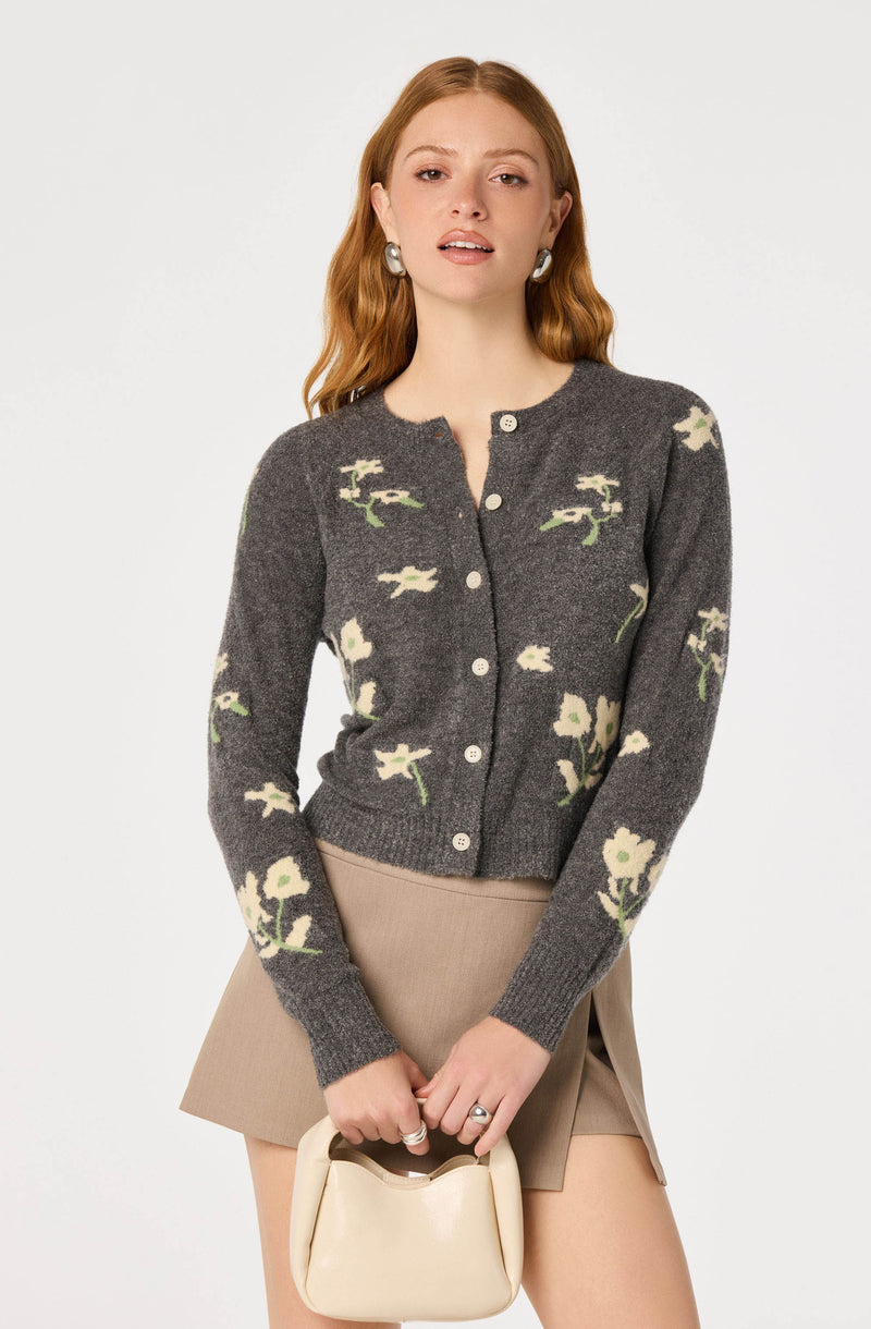 Woman wearing a dark gray floral patterned cardigan with white buttons, beige skirt, and holding a cream-colored handbag.