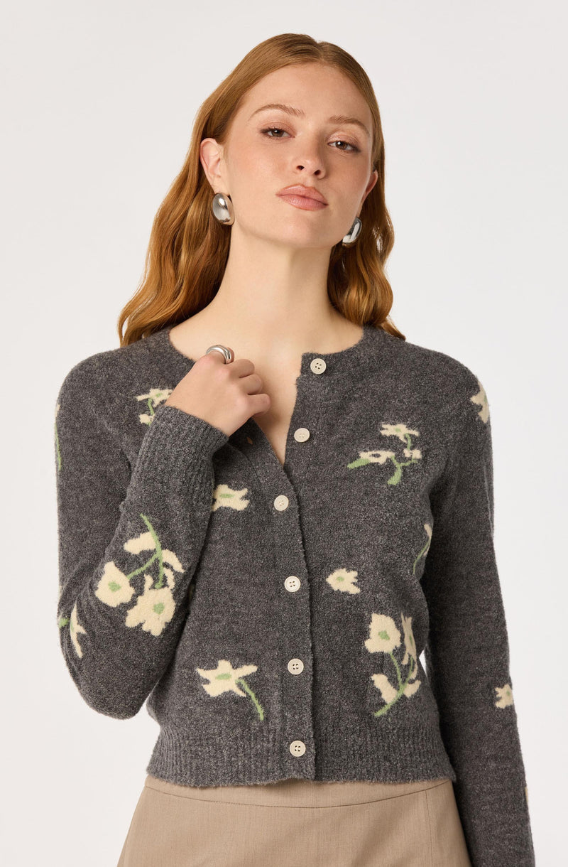 Woman wearing a gray cardigan with white floral patterns and beige pants.