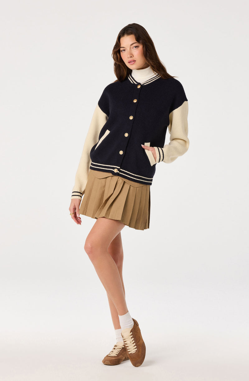 Girl wearing a navy and cream varsity-style cardigan with beige pleated skirt and brown sneakers.