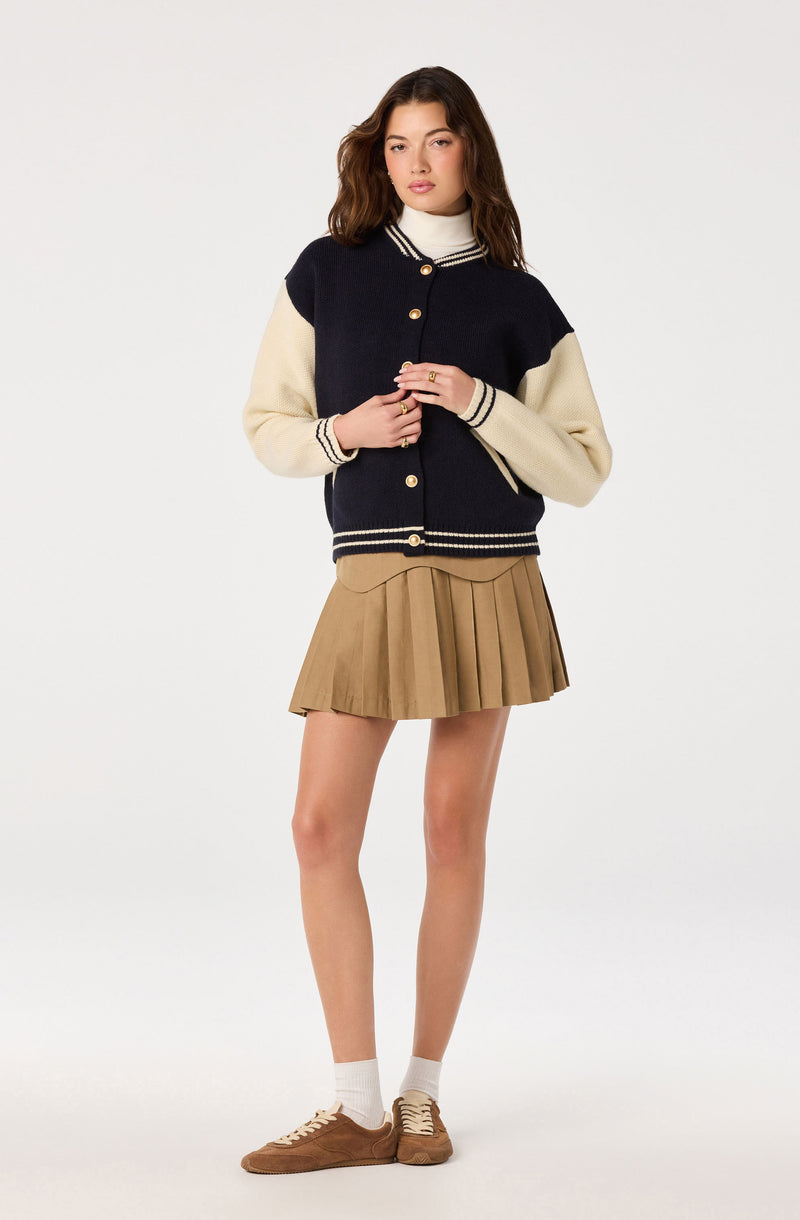 Young woman in a navy and cream varsity jacket, beige pleated skirt, white socks, and brown sneakers.