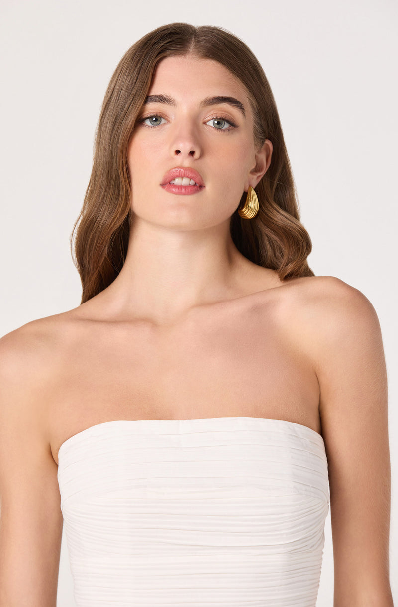 Woman in a strapless white dress with wavy brown hair and gold earrings.