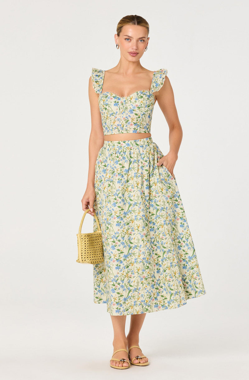 Woman in floral crop top and matching wide-leg skirt, holding a yellow woven handbag.
