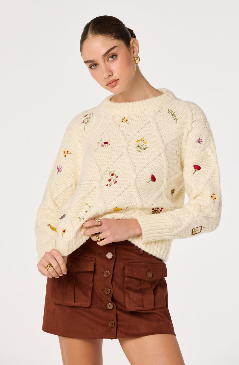 Woman wearing a cream knit sweater with embroidered flowers and a brown button-front skirt.