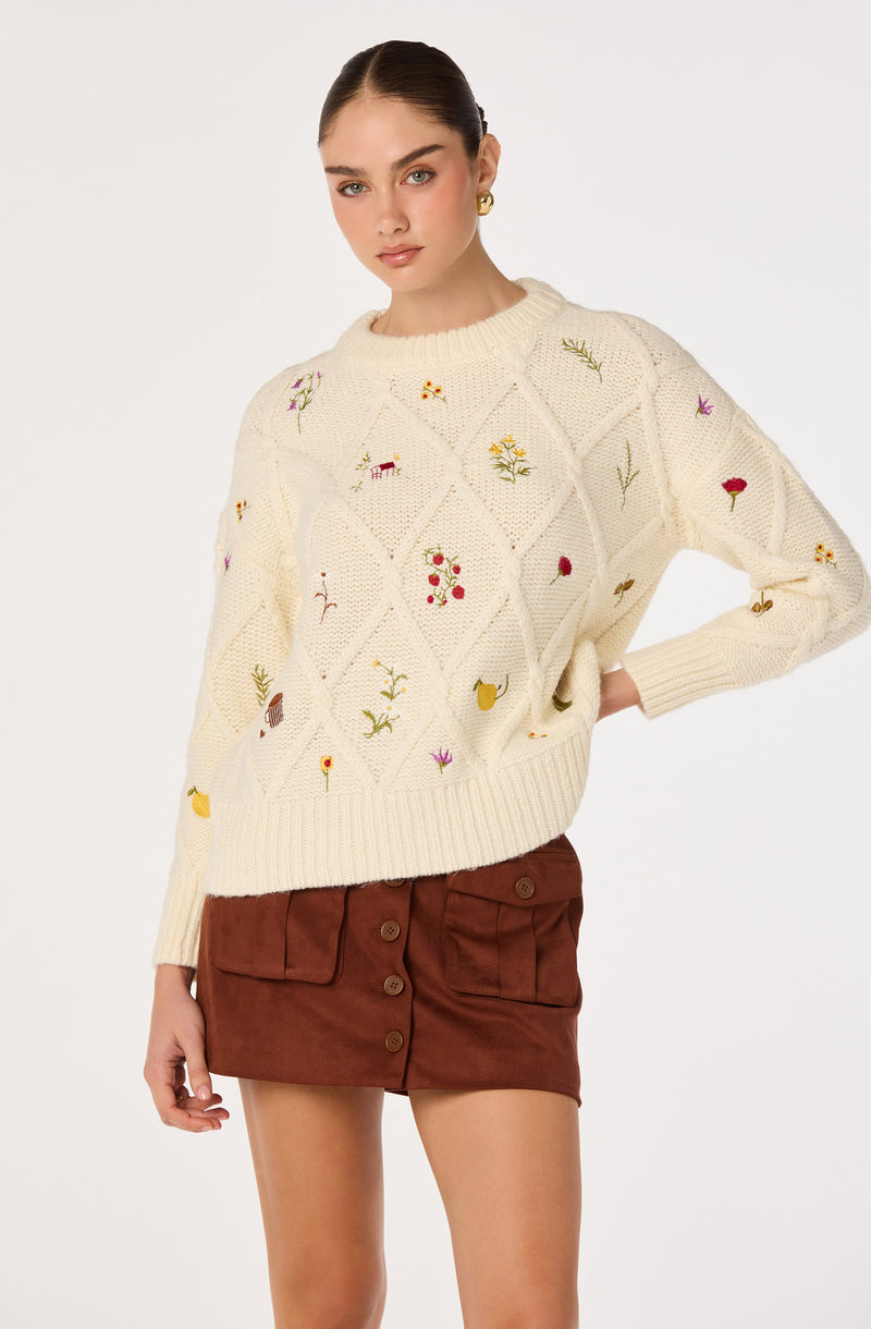 Cream knit sweater with colorful embroidered flowers and leaves, paired with a brown button-front skirt.
