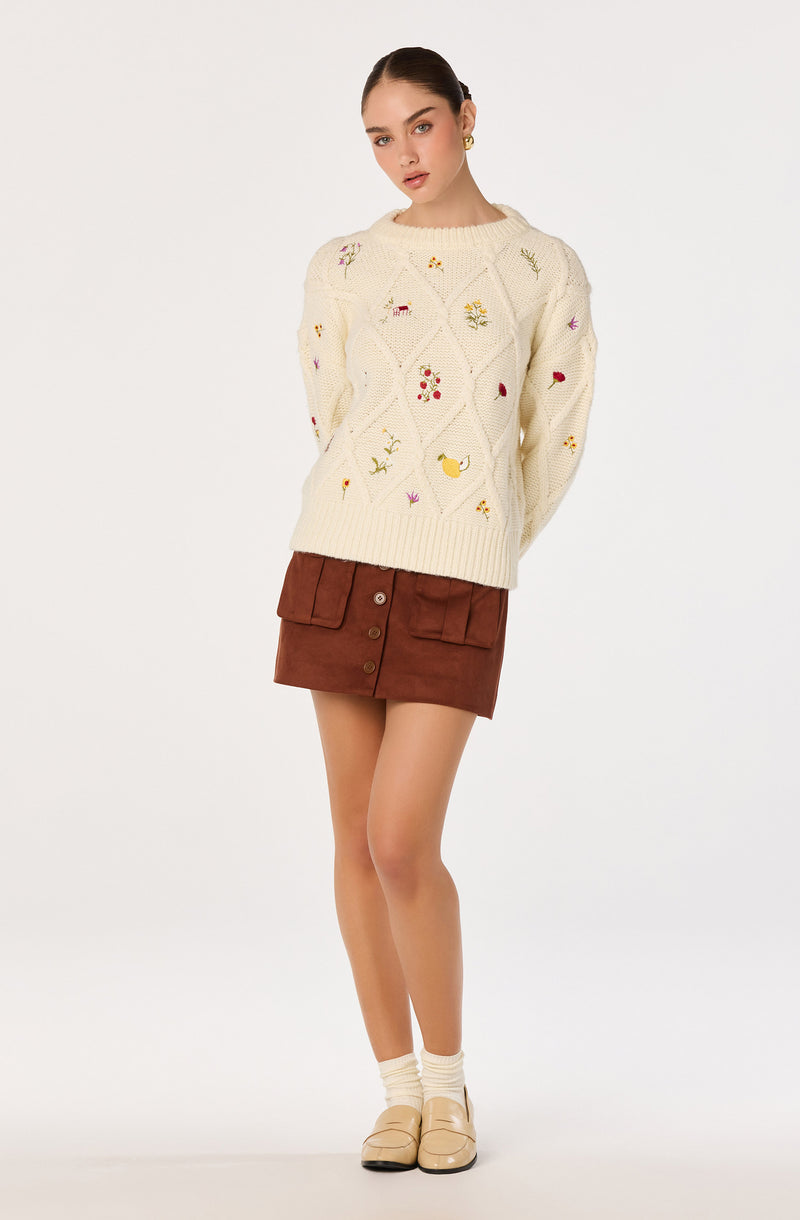 Woman wearing a cream embroidered sweater, brown skirt, cream socks, and beige shoes.