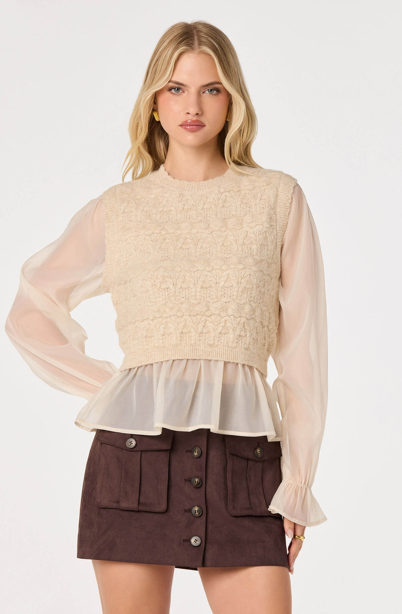 Woman wearing a cream knit vest over a sheer beige blouse with ruffled cuffs and hem.