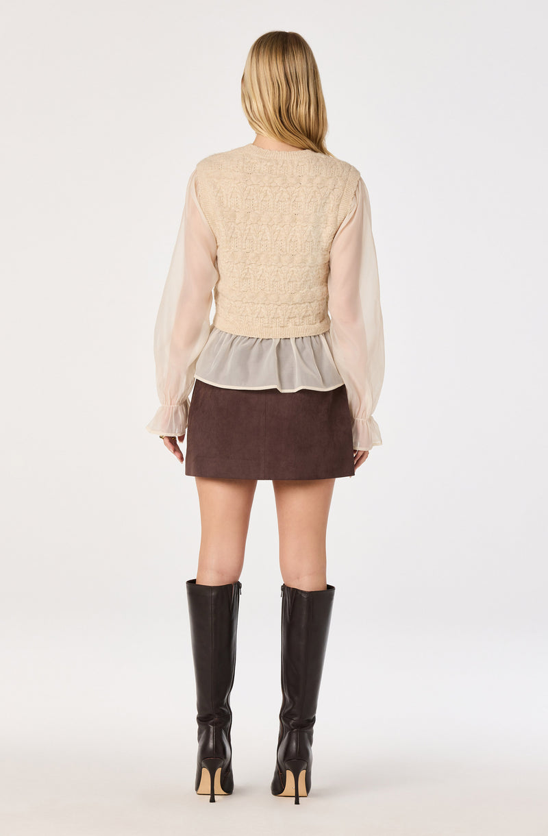 Woman wearing a beige knit vest over a sheer, long-sleeve blouse with ruffled cuffs, paired with a dark brown skirt and knee-high black boots.