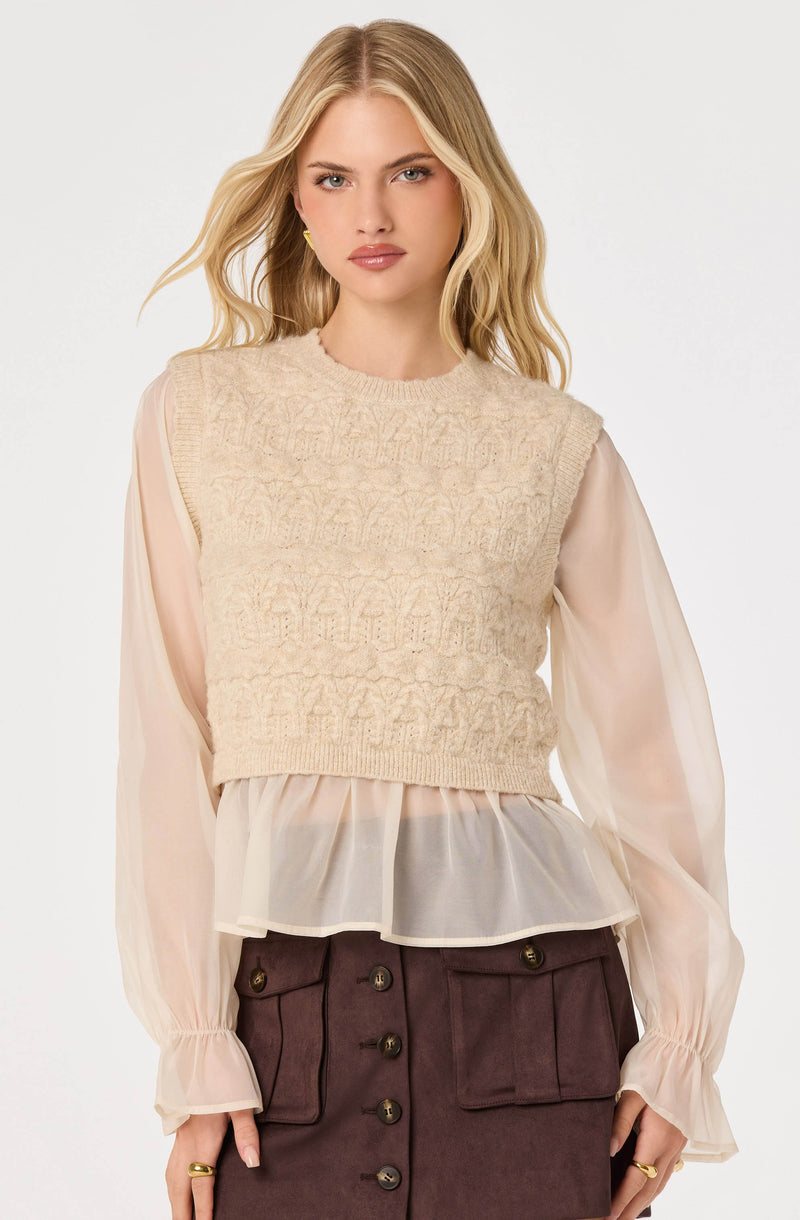 Beige knit sleeveless sweater layered over a sheer, light pink blouse with long puffed sleeves.