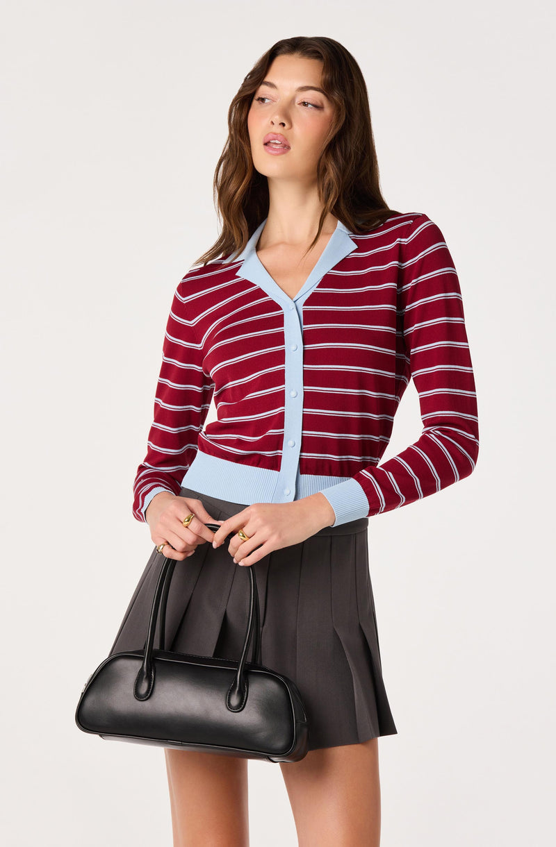 Woman in a red and white striped cardigan with light blue trim, paired with a dark gray pleated skirt, holding a black handbag.