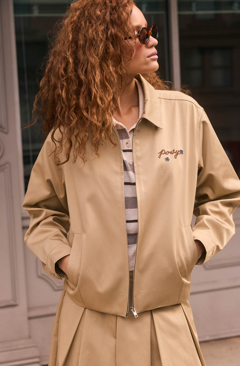 Woman in beige jacket with embroidered text, striped shirt underneath, and sunglasses.
