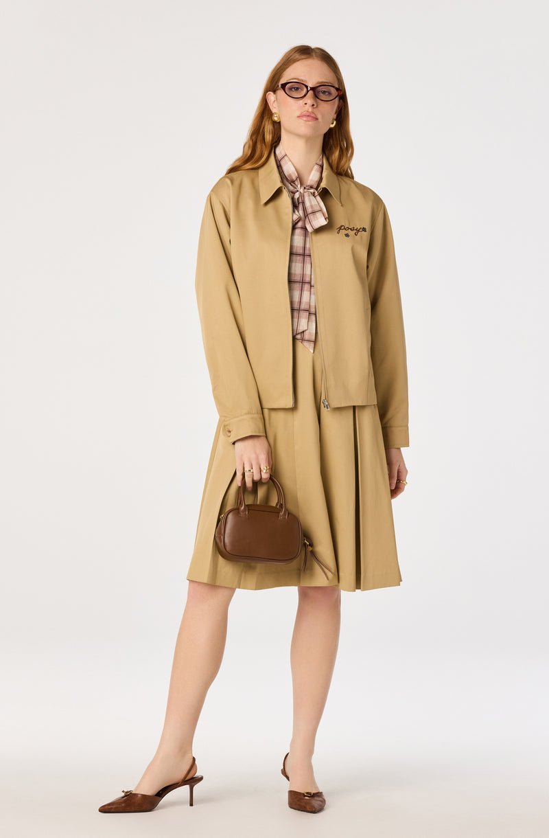 Woman in beige jacket, plaid shirt, pleated skirt, brown heels, and glasses, holding a brown handbag.