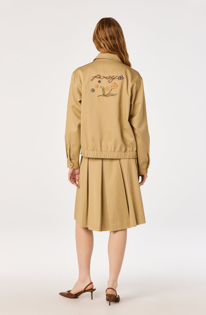 Beige jacket with embroidered floral design and text on the back, paired with a beige pleated skirt and brown heels.