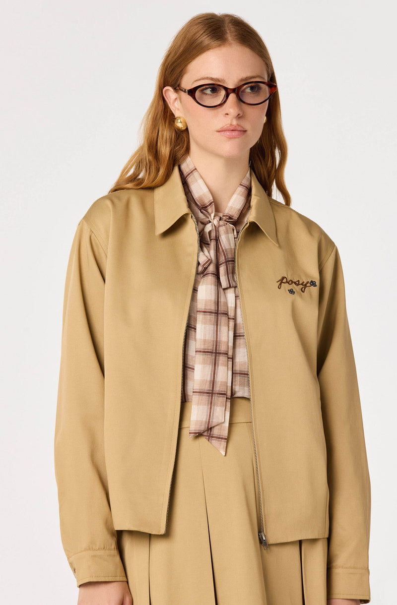 Woman wearing a beige zip-up jacket with embroidered logo, beige pleated skirt, plaid scarf, and glasses.