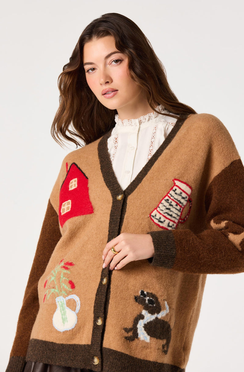 Woman wearing a large, colorful knit cardigan with house, jar, dog, and plant appliqué designs.