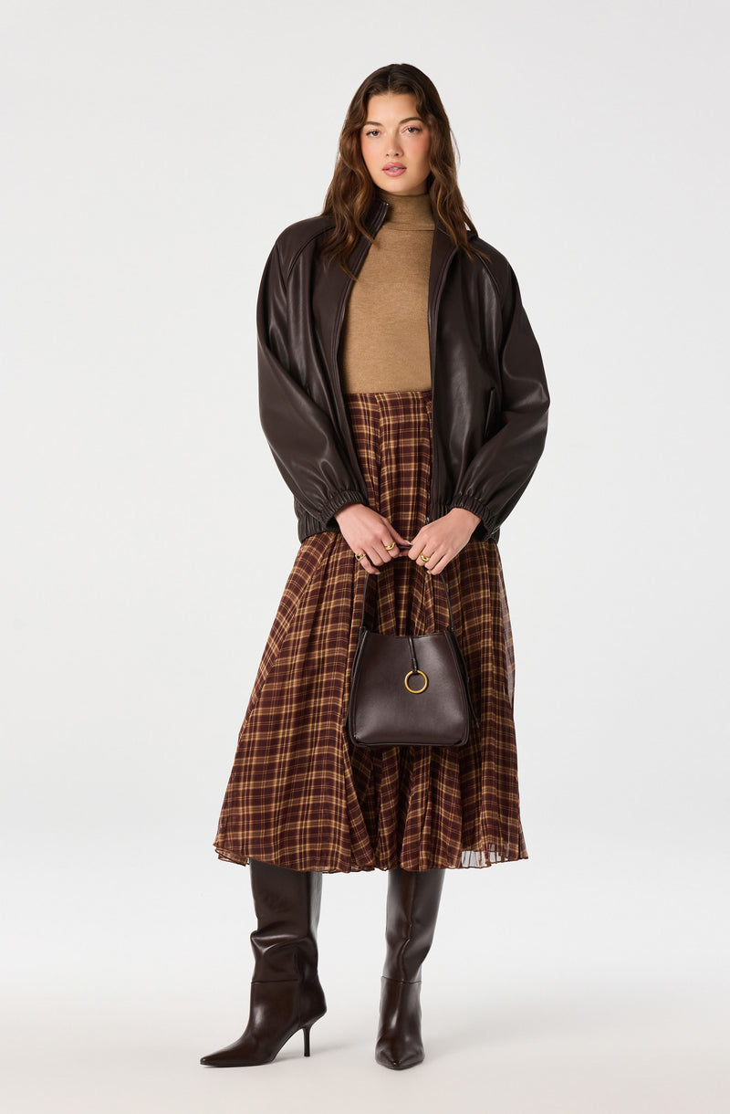 Woman wearing a brown leather jacket, tan turtleneck, plaid A-line skirt, and knee-high leather boots, holding a black handbag.