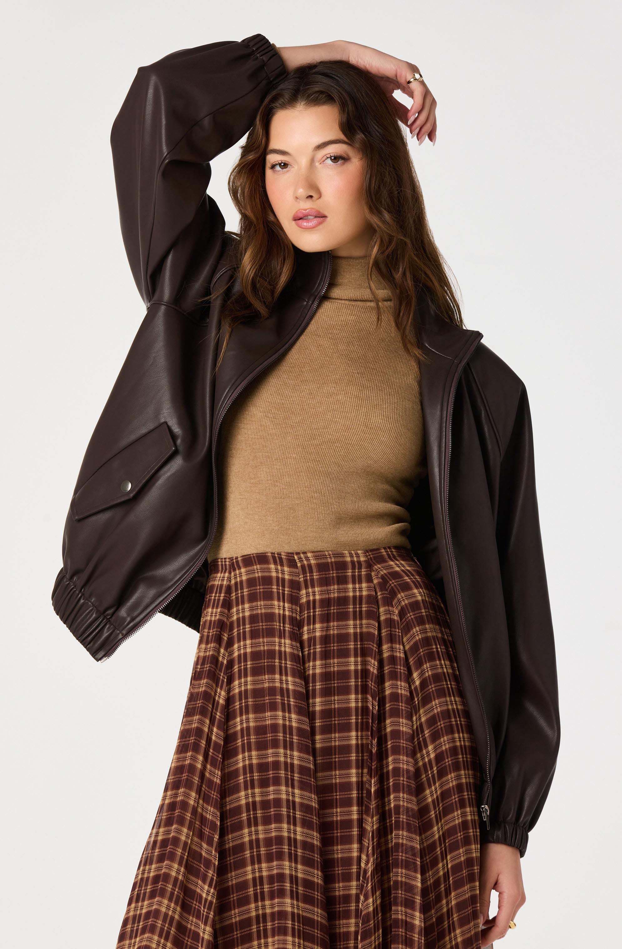 Woman wearing a brown leather jacket, beige turtleneck, and a brown plaid skirt.