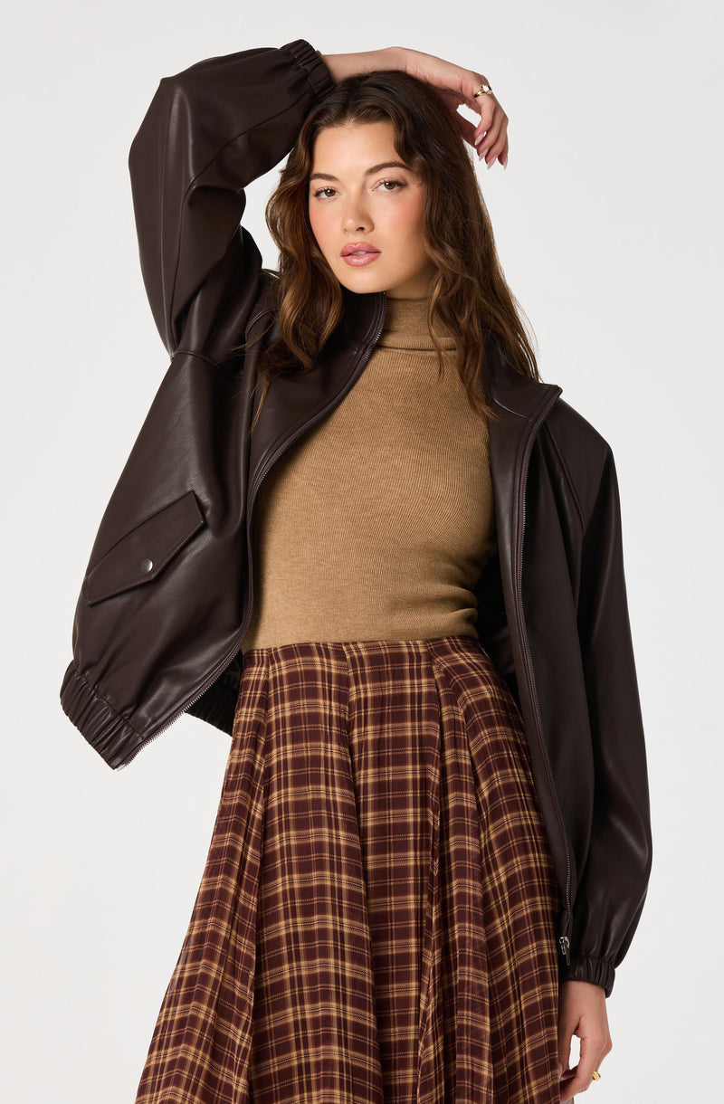 Woman wearing a brown leather jacket, beige turtleneck, and a brown plaid skirt.