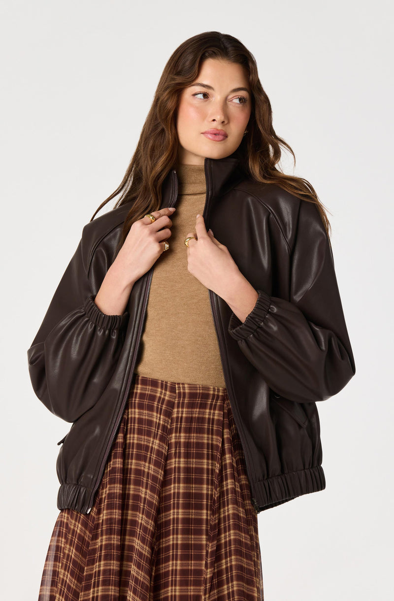 Woman wearing a dark brown leather jacket over a tan turtleneck, paired with a plaid skirt.