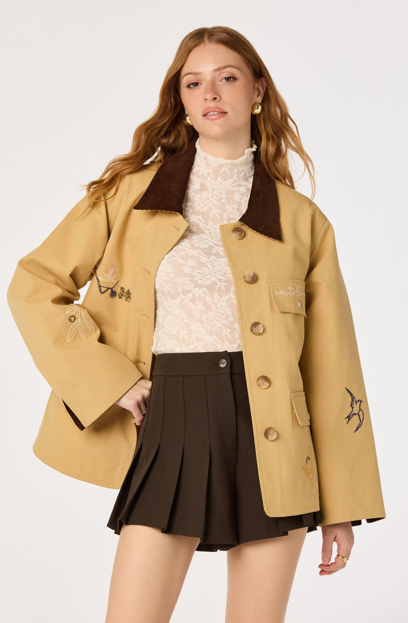 Model wearing a beige jacket with embroidered designs, a white lace top, and a black pleated skirt.