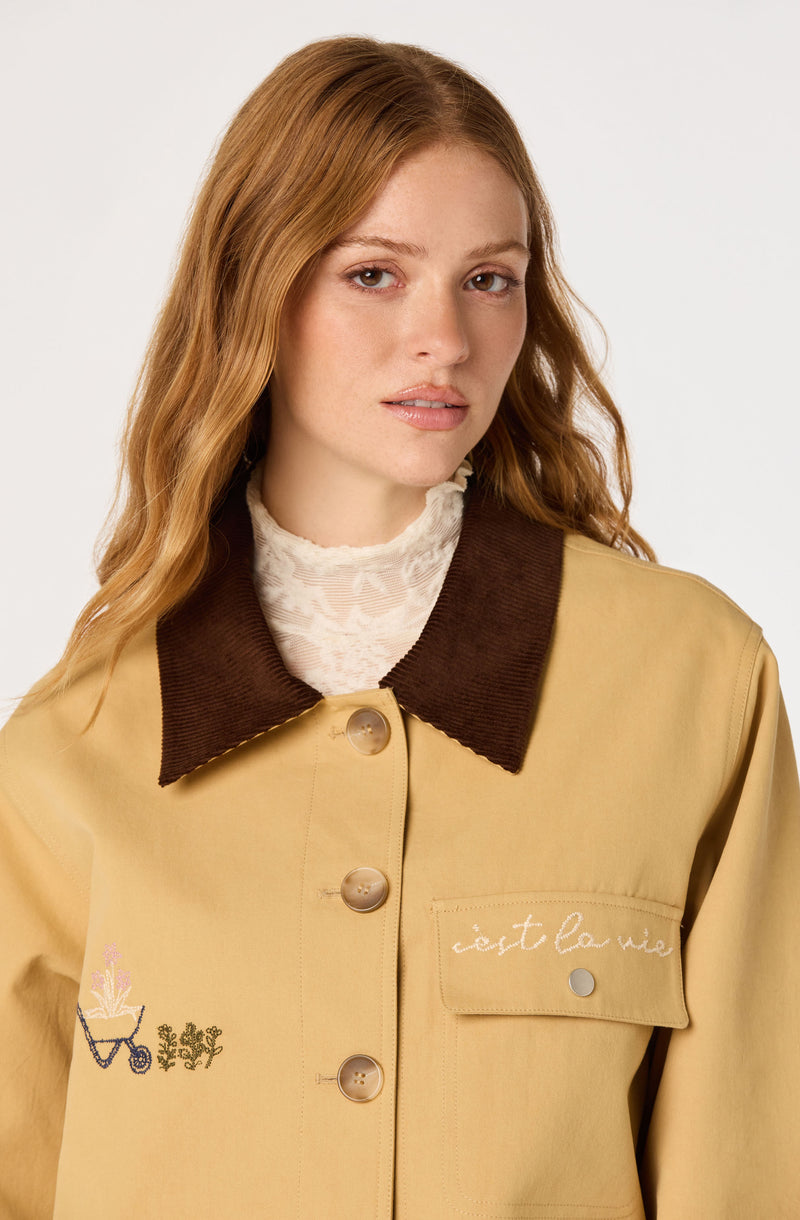 Woman in beige jacket with brown collar, embroidered and button details, over a lace turtleneck.